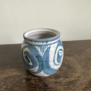May include: A small, round ceramic vase with a wide opening. The vase features a blue and white swirl design. The rim is a darker blue, and it rests on a wooden surface.