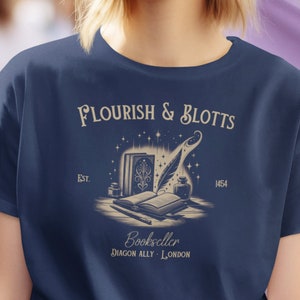 May include: A navy blue t-shirt with a vintage-style illustration of a quill pen, books, and a bottle of ink. The text "Flourish & Blotts" is above the illustration, and "Bookseller Diagon Alley - London" is below. The text "Est. 1454" is on the right side of the illustration.