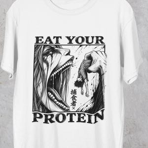May include: A white short-sleeved t-shirt featuring a black and white graphic print of a large open-mouthed creature holding a small figure. The text EAT YOUR PROTEIN is printed in bold black letters above and below the central illustration.
