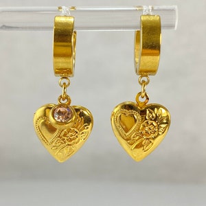 Peut inclure: Une paire de boucles d'oreilles créoles en or avec un pendentif en forme de cœur. Chaque pendentif en forme de cœur présente un motif floral détaillé et un petit strass rose au centre.