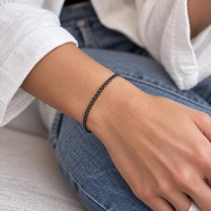 May include: A thin, dark-toned metal chain bracelet featuring a classic curb link design. The bracelet is constructed with small, uniform interlocking links that form a continuous, flexible band, showcasing a sleek, matte black finish throughout.