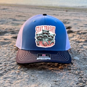 Op de afbeelding: Een blauwe en zwarte trucker-pet met een wit mesh achterpaneel. De voorkant van de pet heeft een afbeelding van vier wasberen met zonnebrillen en de tekst "Stay Trashy". De pet is gemaakt door Richardson 112.