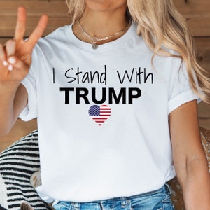 May include: White t-shirt with the text "I Stand With TRUMP" in black lettering and an American flag heart.