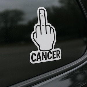 May include: White vinyl sticker depicting a hand giving the middle finger, with the word "CANCER" in a black, bold font below. The sticker is affixed to a car window, with a blurred background.
