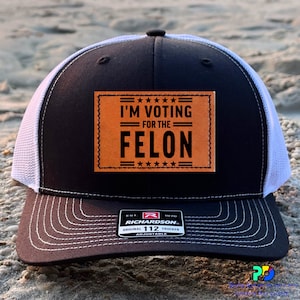 May include: Black and white trucker hat with a brown leather patch that says "I'm Voting for the Felon" with five stars above and below the text. The hat has a white mesh back and a Richardson 112 label.