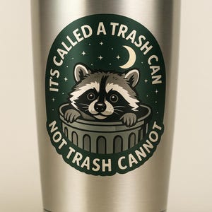 May include: A stainless steel tumbler with a green and white graphic of a raccoon peeking out of a trash can. The text on the tumbler reads "It's called a trash can, not trash cannot."