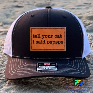May include: Black and white trucker hat with a brown leather patch that says "tell your cat i said pspsps". The hat has a white mesh back and a black front with a white stitching detail. The hat is adjustable and has a Richardson 112 label.
