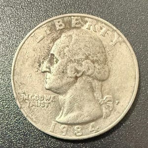 Può includere: Una moneta da un quarto di dollaro statunitense in argento del 1984. La moneta presenta il profilo di una persona con la scritta "LIBERTY" sopra e "IN GOD WE TRUST" sotto. L'anno "1984" è visibile.