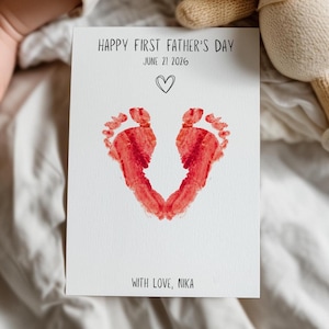 May include: A white card with red footprints forming a heart shape. The card reads "HAPPY FIRST FATHER'S DAY JUNE 21 2026" with a heart symbol and "WITH LOVE, NIKA". A baby's arm and a stuffed animal are in the background.