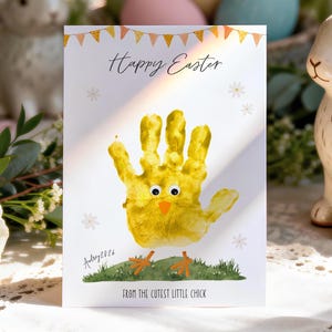 Op de afbeelding: Een witte Paas wenskaart met de tekst "Happy Easter" en een gele handafdruk kuiken illustratie. De kaart bevat ook de tekst "From the cutest little chick" en de naam "Aubrey 2026".