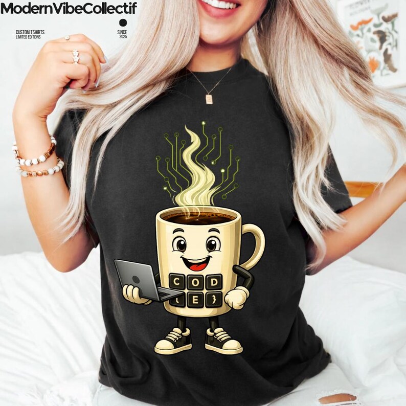Pu&ograve; includere: Una t-shirt nera con la grafica di una tazza di caff&egrave; sorridente che tiene in mano un portatile. La tazza ha un corpo in stile tastiera con il testo "CODE" e "}" stampato sopra, e vapore con un motivo a circuito stampato.