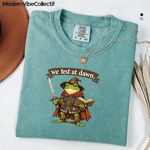 Może przedstawiać: Teal Comfort Colors t-shirt z kreskówkową żabą ubraną jako czarodziej, trzymającą miecz i książkę. Tekst "we test at dawn" znajduje się nad żabą. Srebrny naszyjnik jest zawieszony na koszulce.