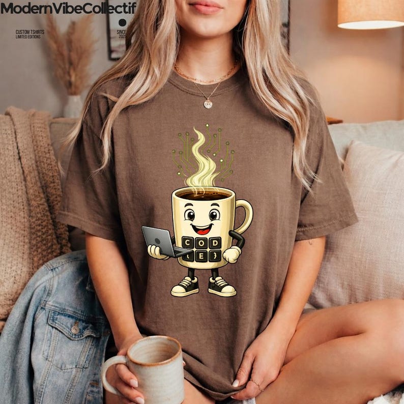 Pu&ograve; includere: Una t-shirt marrone a maniche corte con la grafica di una tazza di caff&egrave; sorridente che tiene in mano un portatile. La tazza ha un corpo simile a una tastiera con la scritta CODE e vapore che sale con un motivo a circuito digitale.