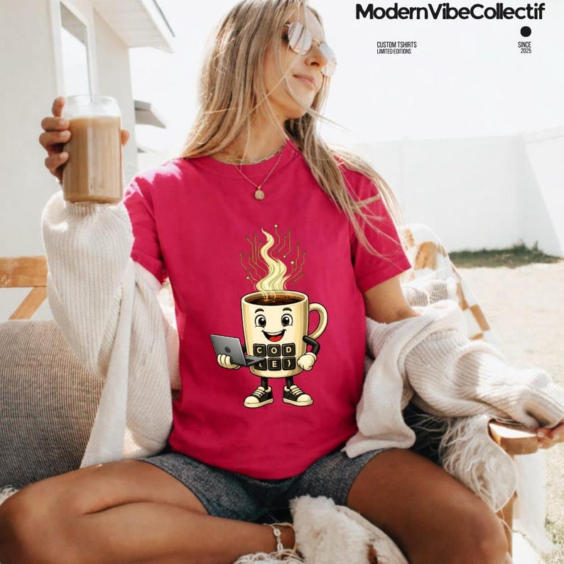 Pu&ograve; includere: Una t-shirt a maniche corte rosa acceso con una grafica a cartoni animati di una tazza da caff&egrave; beige con volto, braccia e gambe. La tazza tiene in mano un piccolo computer portatile e presenta un tastierino frontale con le lettere C, O, D, E.