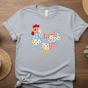 Könnte beinhalten: Hellgraues T-Shirt mit einem Patchwork-Hühner-Design. Das Huhn hat einen roten Kamm, einen gelben Schnabel und Füße sowie einen Körper aus blau, rot und weiß gemusterten Stoffstücken. Das Shirt wird auf einer Holzoberfläche präsentiert.