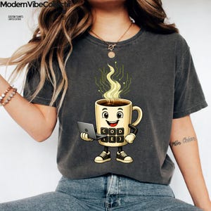 Pu&ograve; includere: Una t-shirt grigio antracite a maniche corte con la grafica di una tazza di caff&egrave; sorridente che tiene in mano un portatile. La tazza presenta un design a tastiera con il testo CODE e parentesi graffe, con vapore a circuito.