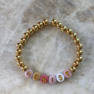 May include: Gold bead bracelet with the word "RESIST" spelled out in pink, white, and beige letter beads.