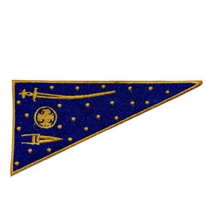 Könnte beinhalten: Ein blaues dreieckiges Wimpel mit einem goldenen gestickten Design. Das Design zeigt ein Schwert, einen Speer und einen Kreis mit einem Vogel darin. Der Hintergrund ist mit kleinen goldenen Punkten bedeckt.