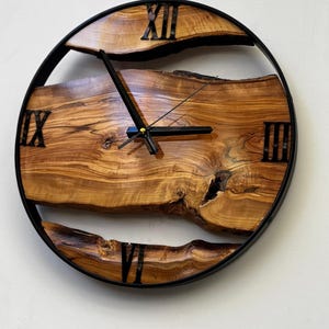 Handmade olive wood wall clock featuring natural live edge wood slabs, black metal frame, Roman numerals, and unique organic wood grain patterns, perfect for rustic, farmhouse, and modern home decor.
