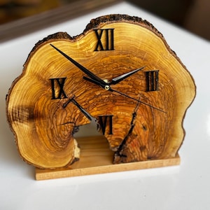 Könnte beinhalten: Eine Holzuhr mit römischen Ziffern auf einem Sockel aus natürlicher Holzscheibe. Das Zifferblatt der Uhr ist aus hellem Holz mit dunkelbraunen Holzmaserungen. Die Zeiger der Uhr sind schwarz und das Zifferblatt ist von einem dunkelbraunen Holzring umgeben.
