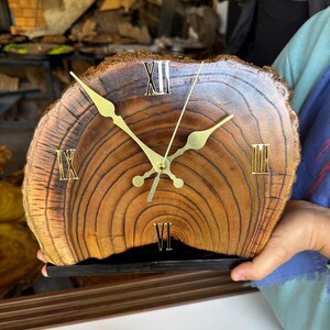 Op de afbeelding: Een houten klok met Romeinse cijfers op een donkerbruine houten voet. De wijzerplaat van de klok is gemaakt van een boomstamschijf, die de natuurlijke houtnerf laat zien.