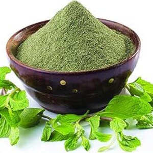 May include: A dark brown ceramic bowl filled with a mound of finely ground green powder, surrounded by several fresh green mint leaves. The bowl features a smooth, rounded rim and a deep, dark finish that contrasts with the vibrant green contents.