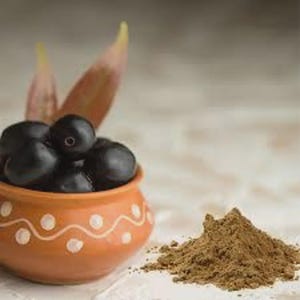 Puede incluir: Un pequeño cuenco de arcilla naranja con puntos y líneas onduladas pintadas en blanco, lleno de frutos jamun de color púrpura oscuro y dos hojas puntiagudas. Junto al cuenco hay un pequeño montón de polvo marrón claro, mostrando la fruta y su forma molida.