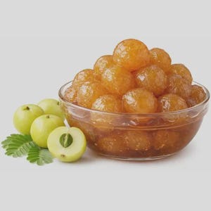 Puede incluir: Un cuenco de cristal transparente lleno de bayas de amla en conserva de color ámbar. Junto al cuenco hay tres frutos de amla enteros de color verde claro y un fruto cortado por la mitad que muestra una semilla, junto a una pequeña rama.