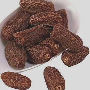 May include: A collection of dried brown dates with wrinkled, textured skin piled inside a white bowl, with two additional dates resting outside the bowl on a plain surface. The dates are oblong in shape and display a deep, dark brown colour.