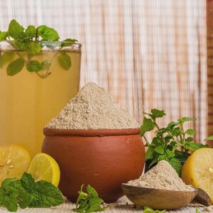 May include: A terracotta clay pot filled with a fine, light beige powder, accompanied by a small wooden scoop containing more of the same powder. Fresh green mint leaves and halved yellow lemons are arranged around the clay pot and scoop.