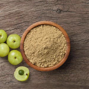 May include: A round wooden bowl filled with fine, light brown herbal powder, positioned next to four whole, smooth, light green amla fruits, one of which is sliced in half to reveal a small, dark green seed in the center of the fruit.