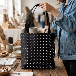 May include: A black tote bag featuring a repeating white polka dot pattern. The bag has two black shoulder straps and is filled with a ceramic mug, a small spiral-bound notebook, and a folded piece of fabric with a blue and orange pattern.