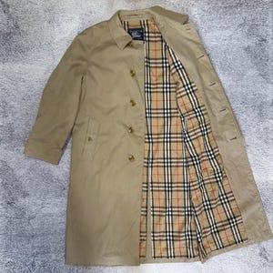 Può includere: Un trench coat beige dal design classico. Il cappotto presenta una chiusura a bottoni, un colletto e una fodera a quadri beige, rossi e neri. Il cappotto è aperto, mostrando la fodera e lo stile.