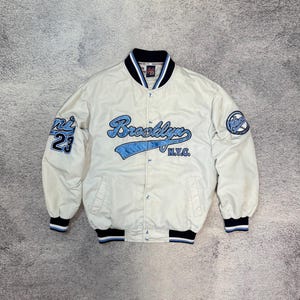 May include: Cream-colored Brooklyn varsity jacket with blue accents. Features the word "Brooklyn" in script across the chest, "N.Y.C." below, and "ni 23" on the left sleeve. The jacket has a button-up front, black and blue striped cuffs, and a circular patch on the right sleeve.