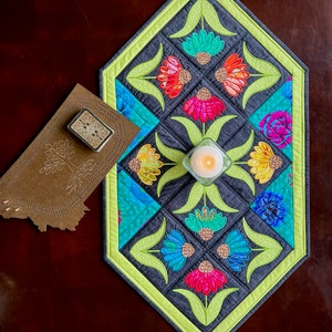 May include: A quilted table runner with a geometric design featuring colorful flowers and green leaves. The runner is made of gray and green fabric with a lime green border.