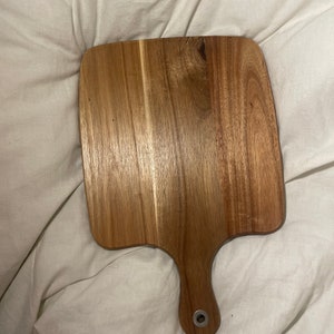 Puede incluir: Una tabla de cortar rectangular de madera con un mango. La tabla tiene un acabado de madera natural y un borde ligeramente redondeado. El mango tiene un anillo de metal.