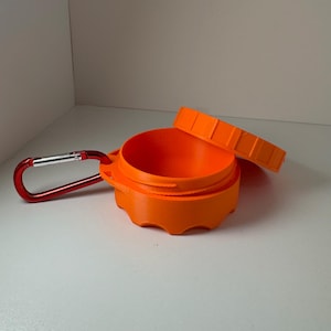 May include: An orange, round container with a matching lid, and a red carabiner clip. The container is open, revealing the interior. The lid has a ridged design. The carabiner is attached to the side of the container.
