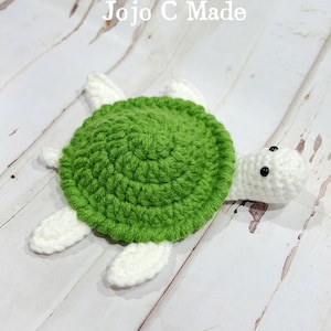 Può includere: Una tartaruga all'uncinetto fatta a mano, verde e bianca. La tartaruga ha un guscio verde, pinne bianche e una testa bianca con occhi neri. Il testo "Jojo C Made" è in lettere bianche sopra la tartaruga.