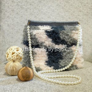 May include: A handmade, fluffy handbag in shades of gray, white, and blue, with a pearl chain strap. The bag has a textured appearance and is rectangular in shape. The text "Handmade By Jojo C." is visible on the bag.