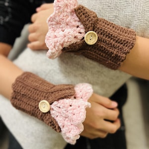 Könnte beinhalten: Ein Paar handgefertigte gehäkelte Handgelenkwärmer. Die Handgelenkbündchen sind dunkelbraun, die gerüschten Kanten sind rosa. Jeder Wärmer hat einen Holzknopfverschluss.