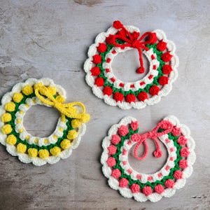 May include: Three handmade crochet wreaths in white, green, red, yellow, and pink. Each wreath has a scalloped edge and a bow. The wreaths are arranged on a light grey surface.