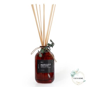 以下が含まれることがあります： 黒いラベルに「No. 4 Marigold Orchid Fragrance Diffuser」と書かれた茶色のガラス瓶。瓶は透明な液体で満たされ、木製の棒が突き出ています。瓶の上部には緑の葉が飾られています。