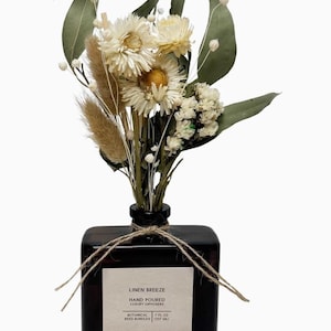 Op de afbeelding: Een donkerbruine vierkante glazen fles met een boeket van gedroogde bloemen en groen. De fles is vastgebonden met touw en heeft een label met de tekst "Linen Breeze". Het boeket bevat witte en gele bloemen en groene bladeren.