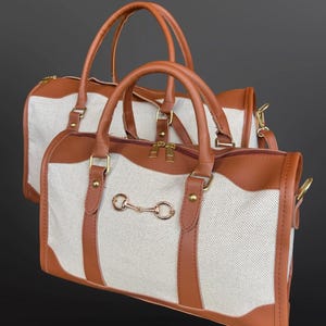 May include: Two duffel bags featuring a cream-colored textured fabric body with brown faux leather trim, handles, and piping. Each bag includes a gold-toned metal horse bit ornament on the front, gold hardware, and a top zipper closure.
