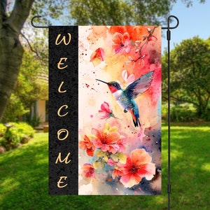May include: A watercolor garden flag with a hummingbird in flight, surrounded by pink and orange flowers. The text "WELCOME" is printed vertically on the left side of the flag.