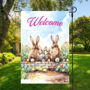 May include: A welcome garden flag with a watercolor design of four bunnies sitting on a brick wall with colorful Easter eggs. The bunnies are looking up at the sky and the text "Welcome" is written in pink letters on the flag.