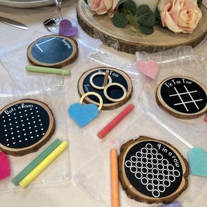 May include: Wooden coasters with chalkboard centres, displaying games such as Tic Tac Toe, Dots and Boxes, and 4 in a Row. Each coaster has a natural wood edge, with colourful chalk sticks and felt heart decorations.