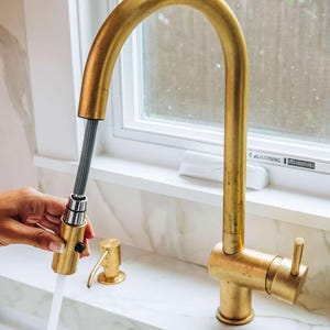 Può includere: Un rubinetto da cucina dorato con uno spruzzino estraibile. Il rubinetto ha un'unica maniglia e un becco ad arco alto. Il rubinetto è montato su un piano di lavoro bianco.