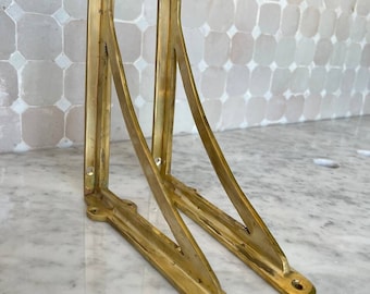 May include: Two gold metal shelf brackets with a triangular shape. The brackets are made of solid metal and have a polished finish.