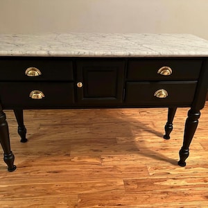 May include: A black console table with a white marble top. The table has four drawers with gold-colored handles and a central cabinet. The legs are black and turned. A stylish piece of furniture.